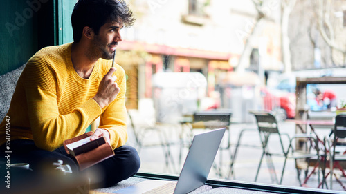 Handsome male creative journalist feeling happy having brilliant idea for new article working on distance job sitting in coffee shop noting text of future reportage into notebook before using laptop