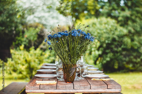 Close up of blue cornflowers in a vase on a dinner table. Blurred background. Midsummer flowers from a meadow. Empty dishes set on a table 
