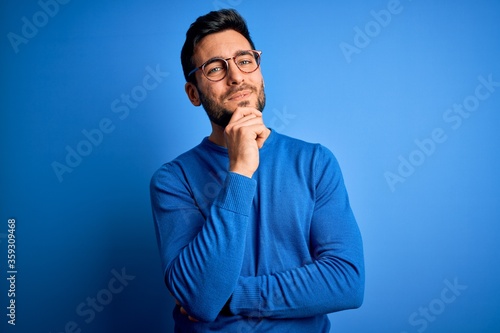 Young handsome man with beard wearing casual sweater and glasses over blue background looking confident at the camera smiling with crossed arms and hand raised on chin. Thinking positive.