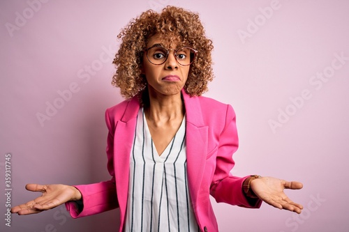 Young african american businesswoman wearing glasses standing over pink background clueless and confused with open arms, no idea concept.