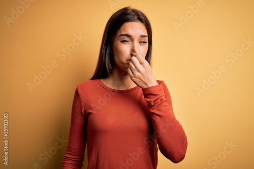 Young beautiful brunette woman wearing casual t-shirt standing over yellow background smelling something stinky and disgusting, intolerable smell, holding breath with fingers on nose. Bad smell