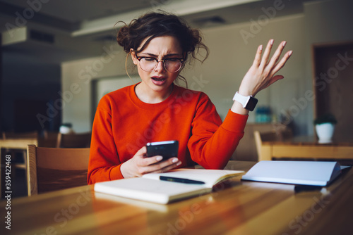 Angry attractive female feeling disappointed receiving bill from banking service checking email box in smartphone connected to wireless internet, emotional hipster girl upset about failure of payment