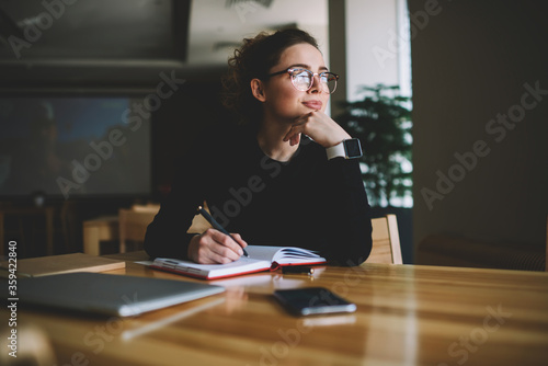 Thoughtful young author businesswoman and thinking on writing interesting article.Woman in stylish eyeglasses with good lenses looking out of window and pondering new plans while noting them