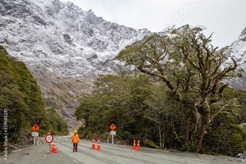 The alpine road is closed to traffic going into Milford Sound, while the traffic leaving Milford Sound travels through the Homer Tunnel. A female road work is in attendance. New Zealand