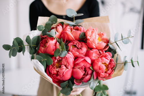 Very nice young woman holding beautiful tender blossoming mono bouquet of fresh coral peony flowers and eucalyptus on the rustic wooden wall background