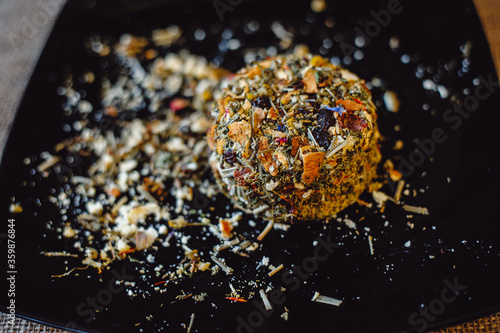 The round head of the French spicy cheese Boulette d'Avesnes sprinkled with different herbs, lies on a black plate. Close-up. Brin d'Amour