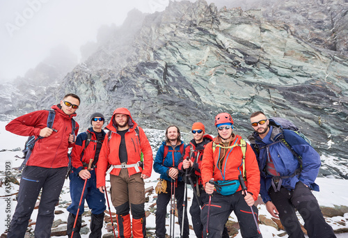 Travelers team in winter jackets and hiking sunglasses posing near huge rocky mountain. Group of backpackers with trekking sticks looking at camera. Concept of travelling, hiking and mountaineering.