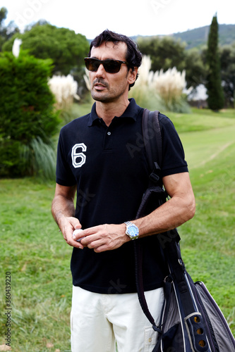 Portrait of stylish golfer man in glasses standing on beautiful golf course looking away, handsome brunette hair man with golf bag on shoulder holding ball in the hands