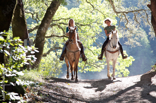 Two women riding horses toward camera with backlit leaves