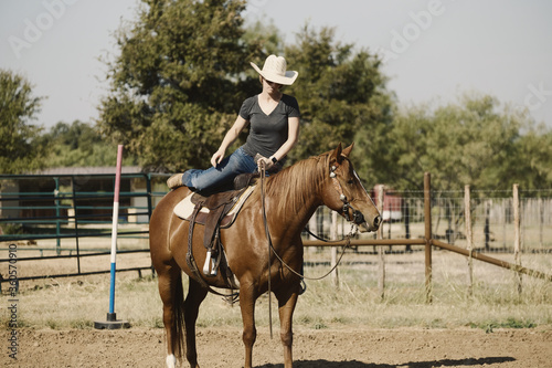 Western lifestyle shows cowgirl mounting in saddle on horse to ride outdoors in cowboy hat.