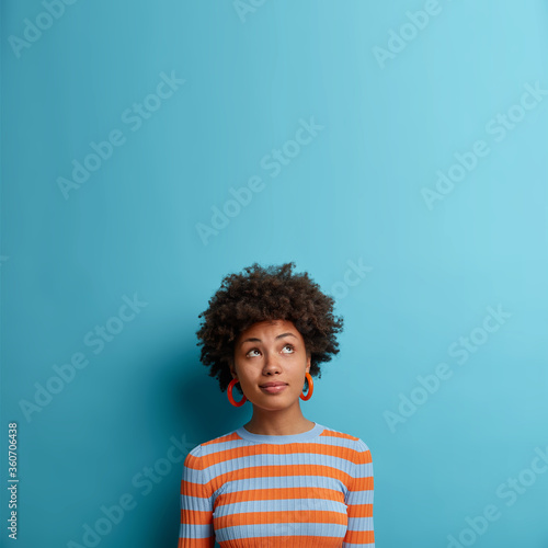 Vertical shot of cute dark skinned woman with Afro hairstyle, looks above with pondering expression, notices something interesting, blank space on blue background. Face expressions and emotions
