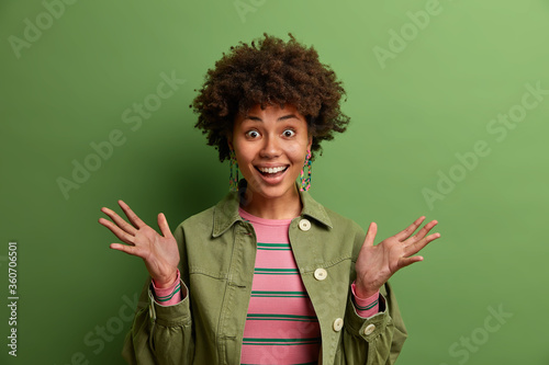 Cheerful young woman raises palms and smiles broadly, has happy reaction on something awesome, dressed in fashionable clothes, poses against green background. People, positive emotions, reaction