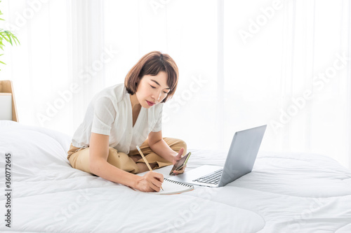 asian business woman work from home via laptop on bed with smartphone and notebook.