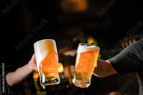 two hands with beer mugs, toasting in celebration, with overflowing foam, dark background and space for writing