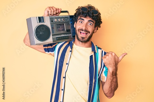 Handsome young man with curly hair and bear holding boombox, listening to music wearing summer look pointing thumb up to the side smiling happy with open mouth