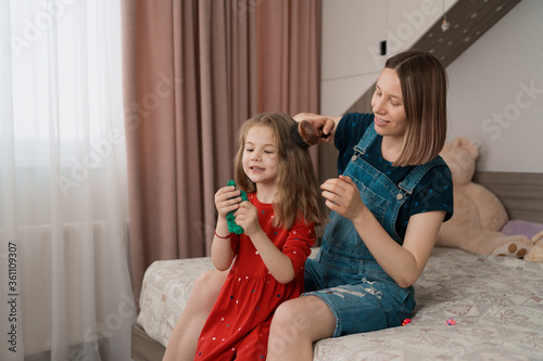 Happy delightful aunt wearing comfy jeans romper, spend time with her cute lovely niece, combing her hair, they are sitting on a bed at home