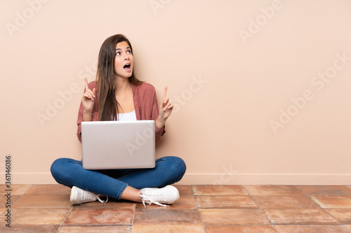 Teenager student girl sitting on the floor with a laptop surprised and pointing up
