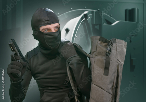 heist and robbery - Hollywood style portrait of man in balaclava mask holding gun in front of security metal vault door in bank or casino heist concept stealing money