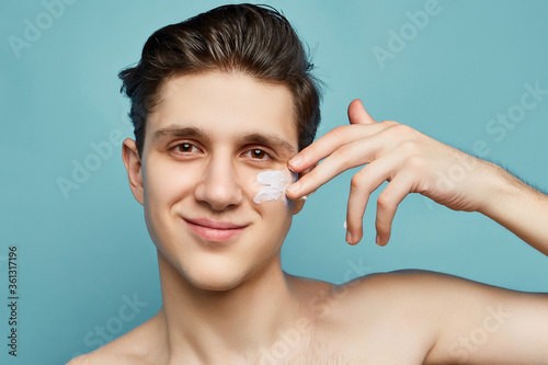 Busy men prefer minimalist, easy and quick facial treatments. A young smiling man is making white brushstrokes under his eye using white creamy mass.