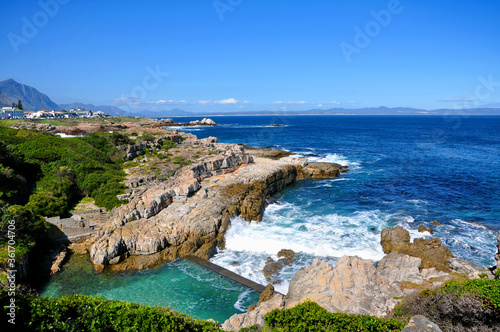 Beautiful Atlantic Ocean coast in Hermanus with waves crashing on the shore and houses on the cliff on sunny day, South Africa