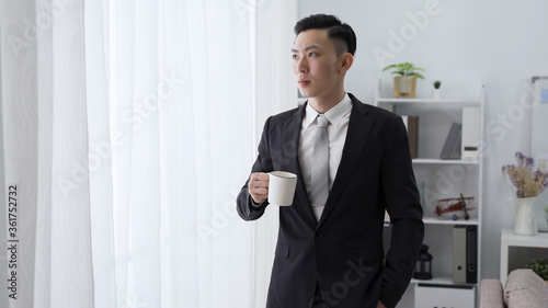 male ceo is sipping morning coffee by window with serious expression. asian man in black suit with hand in pocket is looking outside in contemplation