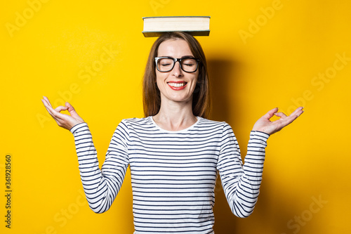 Young woman meditates with closed eyes holds a book on her head on a yellow background.