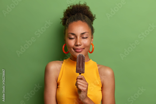 Pleased attractive woman poses for vacation photo shoot, holds delicious chocolate ice cream, closes eyes and imagines pleasant flavor, has happy time wears yellow t shirt isolated on green background