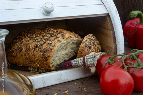 Fresh baked bread with seeds (sesame, sunflower seeds, linen) in white wooden bread box. Breadbox with cutted bread, tomatoes, olive oil in glass bottle and red bell peppers on the wooden background.