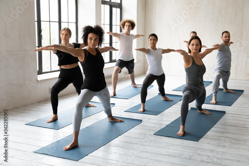Strong sporty african american female instructor giving yoga class in modern studio, standing indoors with young happy mixed race people in warrior II second pose, enjoying working out together.