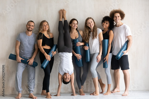 Sportive fit smiling indian ethnicity woman standing upside down on hands near wall with young happy excited multiracial people in activewear holding floor mats, warming up before training indoors.