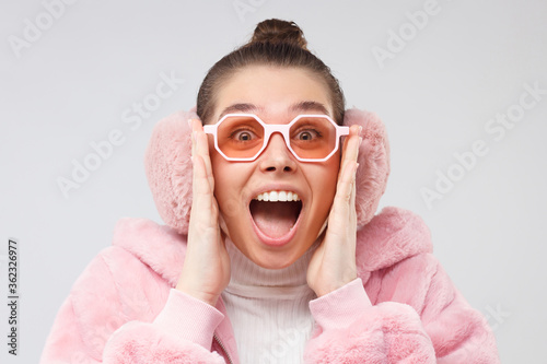 Young emotional trendy girl dressed in pink fur coat, wearing earmuffs and glasses, shouting wow with surprise, isolated on gray background