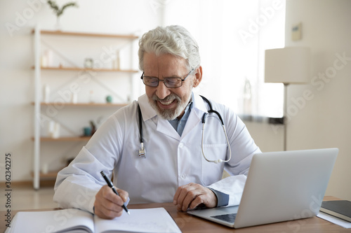 Smiling old male doctor in glasses and white uniform sit at desk in hospital work on laptop write in journal, happy mature senior man physician fill patient medical history anamnesis in paper register