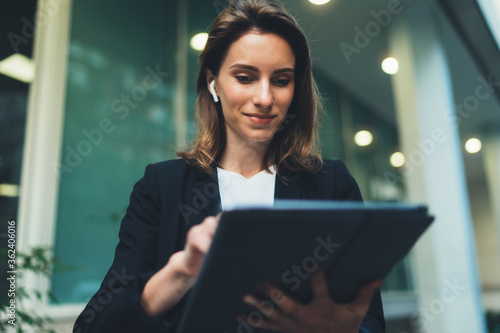 Successful female banker using tablet and wireless earphones outdoors near his office background lights, portrait young woman professional manager working on touch pad near skyscraper in evening city