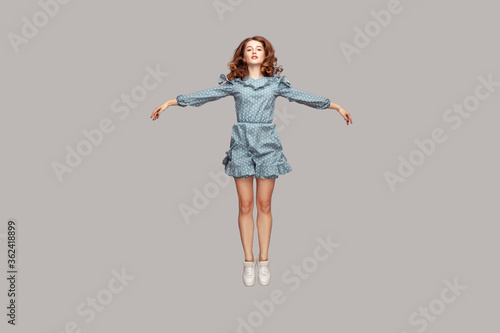 Full length happy calm pretty girl in vintage ruffle dress levitating hovering in mid-air with raised hands as wings, jumping trampoline or flying up. indoor studio shot isolated on gray background