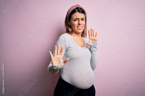 Young beautiful brunette woman pregnant expecting baby over isolated pink background disgusted expression, displeased and fearful doing disgust face because aversion reaction. With hands raised