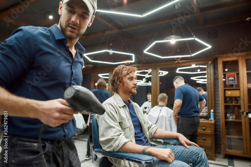 Getting new haircut. Young male barber going to dry a hair of handsome redhead guy sitting in barber chair in front of the mirror in the modern barber shop