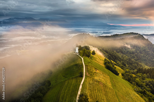 Jamnik, Slovenia - Aerial drone view of a foggy summer sunrise at Jamnik St.Primoz church. The fog gently goes around the small chapel with colorful sky and Julian Alps at background