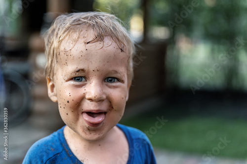 Cute adorable blond caucasian little happy toddler boy portrait with messy mus spots on face after playing watering garden at yard or countyside farm. Childhood happiness summertime country concept