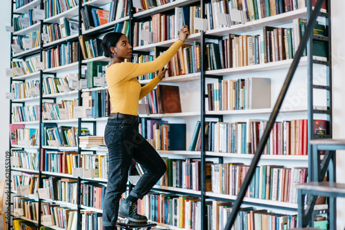 African american female librarian dressed in casual wear choosing literature book for exam preparation.Dark skinned hipster student pulling out book from bookshelf spending time in library