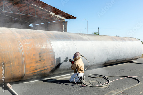 View of the manuel sandblasting to the large pipe. Abrasive blasting more commonly known as sandblasting is the operation of forcibly propelling a stream of abrasive material against a surface.