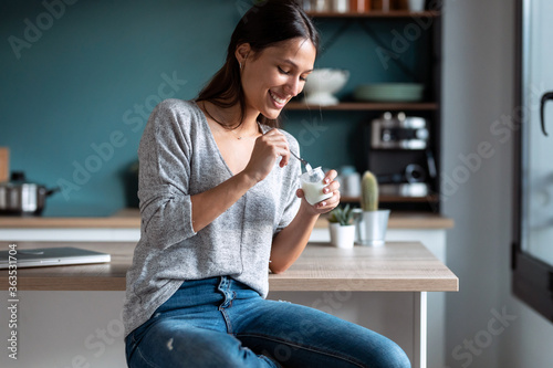 Smiling young woman eating yogurt while sitting on stool in the kitchen at home.