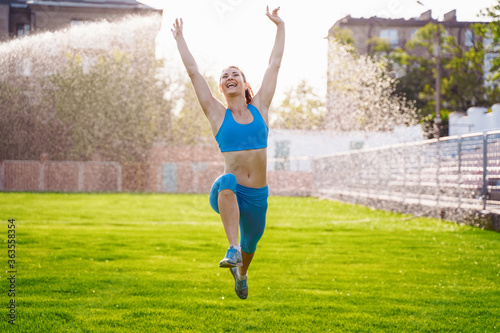 Beautiful brunette woman athlete, wearing top and tight leggings, running through a sports stadium covered with grass. Performing an exercise jump. Happily bouncing from the good news. Hands up