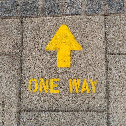 Bright yellow one way arrow sign spray painted onto street to direct pedestrians which way to walk around the town centre during Corona Virus pandemic in the United Kingdom