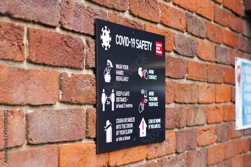 COVID 19 safety information sign for shoppers mounted on brick wall leading to town centre shopping area with shallow depth of field