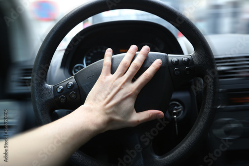 A panicking hand pushing the steering wheel of a car whilst driving. Honking the horn can be used to avoid an accident in a dangerous situation. The car is clearly driving fast, or is even speeding.