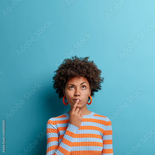 Vertical shot of thoughtful dark skinned woman has idea in mind, keeps finger on lips, looks pensively above, tries to decide how to act, poses against blue background, blank copy space above