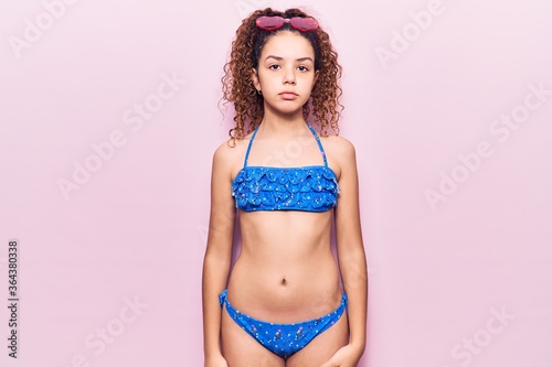 Beautiful kid girl with curly hair wearing bikini and sunglasses relaxed with serious expression on face. simple and natural looking at the camera.