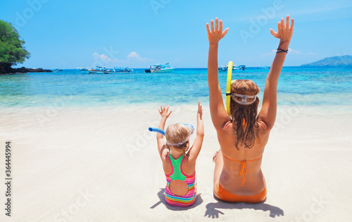 Happy family - mother, baby girl in snorkeling mask sit on white sand beach of tropical island. Look at scenic sea lagoon view. Travel adventure, swimming activity on summer holiday cruise with kids.