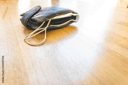 Black bulky wallet with facial mask in it laying on the light wooden textured surface with blank space. Concept of safety regulations in restaurants and public places during global pandemic.