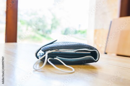 Balck bulky wallet with facial mask in it laying on the light wooden textured surface with blank space. Concept of safety regulations in restaurants and public places during global pandemic.